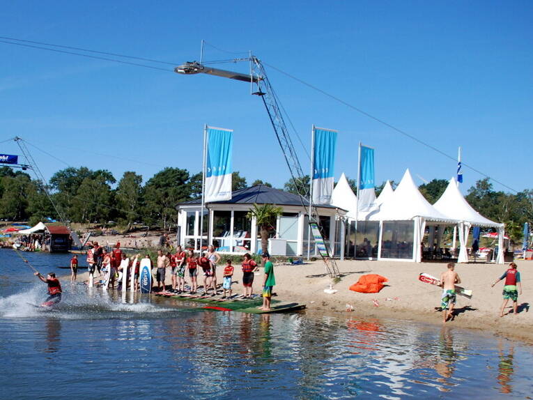 Am Ufer des Blauen Sees hat sich eine Schlange von Menschen gebildet, die auf Wakboard oder Wasserskiern über das Wasser gezogen werden wollen. Am Ufer des Blauen Sees hat sich eine Schlange von Menschen gebildet, die auf Wakboard oder Wasserskiern über das Wasser gezogen werden wollen.