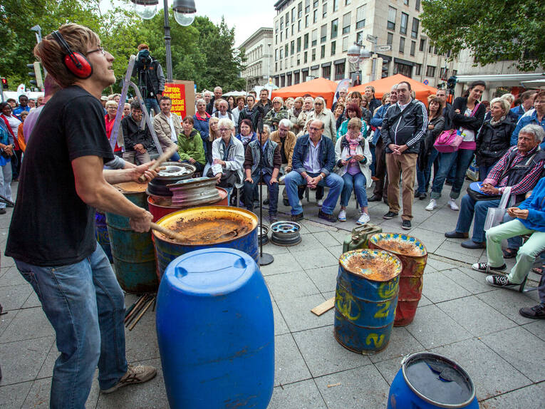 Ein Musiker trommelt auf einer blauen Tonne aus Plastik, vor und neben ihm stehen weitere Fässer, diese sind aber zum größten Teil aus Metall. Ein Musiker trommelt auf einer blauen Tonne aus Plastik, vor und neben ihm stehen weitere Fässer, diese sind aber zum größten Teil aus Metall.