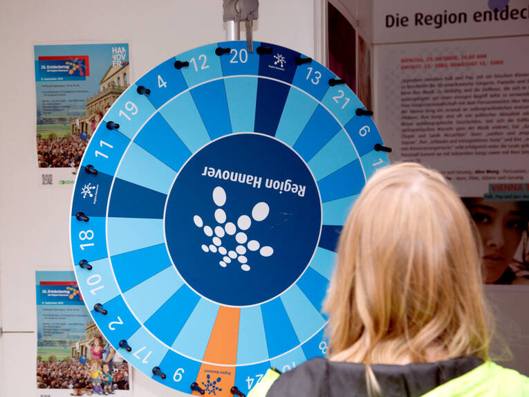 Ein Mädchen steht vor einem Glücksrad. Die Felder des Glücksrades sind überwiegend blau, in der Mitte ist das Punktelogo der Region Hannover. Ein Mädchen steht vor einem Glücksrad. Die Felder des Glücksrades sind überwiegend blau, in der Mitte ist das Punktelogo der Region Hannover.