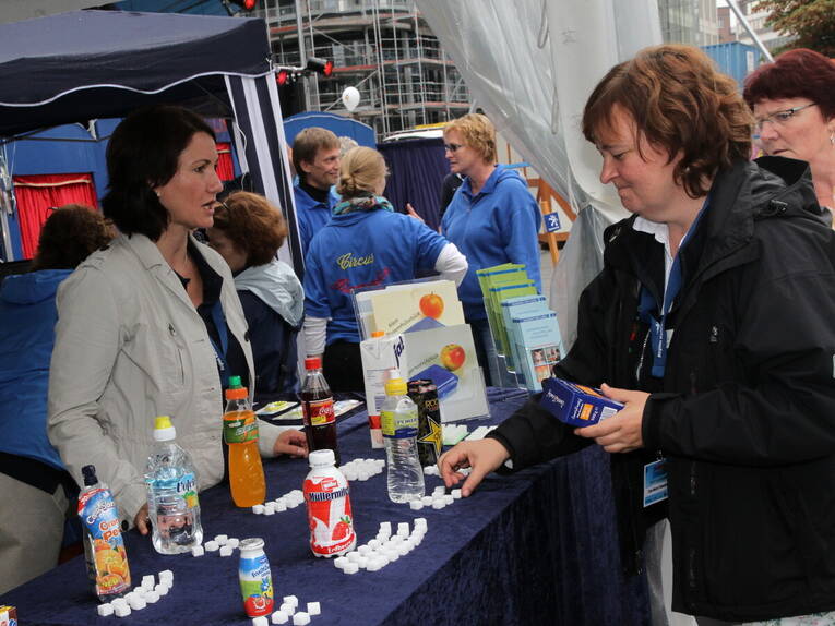 Eine Mitarbeiterin der Region Hannover steht hinter einem Stand beim Entdeckerfest. Auf einem Tresen stehen verschiedene Getränk,e vor jedem Getränk liegen Zuckerwürfel. Eine zweite Mitarbeiterin steht vor dem Tresen und platziert weitere Zuckerwürfel. Eine Mitarbeiterin der Region Hannover steht hinter einem Stand beim Entdeckerfest. Auf einem Tresen stehen verschiedene Getränk,e vor jedem Getränk liegen Zuckerwürfel. Eine zweite Mitarbeiterin steht vor dem Tresen und platziert weitere Zuckerwürfel.