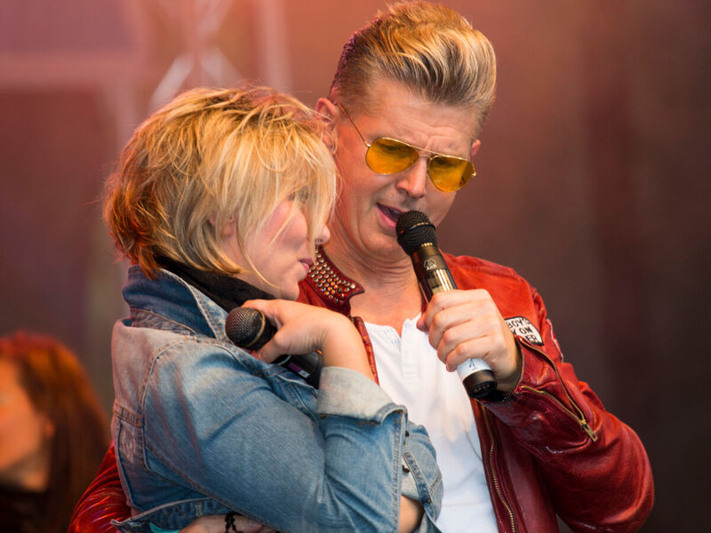 Eine blonde Frau und ein blonder Mann stehen auf einer Bühne, er nimmt sie in den Arm und singt in ein Mikrofon. Eine blonde Frau und ein blonder Mann stehen auf einer Bühne, er nimmt sie in den Arm und singt in ein Mikrofon.