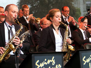 Mehrere Künstler und eine Künstlerin stehen oder sitzen auf einer Bühne und spielen Saxophon oder Trompete. Mehrere Künstler und eine Künstlerin stehen oder sitzen auf einer Bühne und spielen Saxophon oder Trompete.