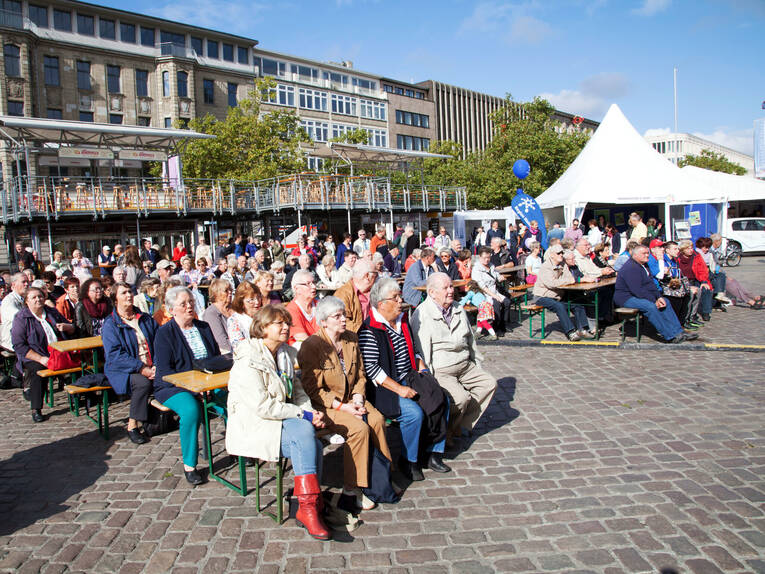 Zuschauerinnen und Zuschauer sitzen auf den Bänken auf dem Opernplatz und lauschen einem Seemannschor. Zuschauerinnen und Zuschauer sitzen auf den Bänken auf dem Opernplatz und lauschen einem Seemannschor.