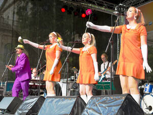 Drei Frauen tragen orangefarbene Kleider und singen in Mikrofone. Daneben steht ein Mann, der ebenfalls in ein Mikrofon singt und einen violetten Anzug trägt. Drei Frauen tragen orangefarbene Kleider und singen in Mikrofone. Daneben steht ein Mann, der ebenfalls in ein Mikrofon singt und einen violetten Anzug trägt.