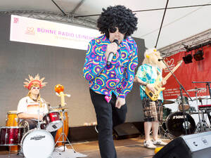Ein Sänger, ein Gitarrist und ein Schlagzeuger spielen auf einer Bühne, sie tragen dabei besonders ausgefallene, bunte Kleidung und Perücken. Ein Sänger, ein Gitarrist und ein Schlagzeuger spielen auf einer Bühne, sie tragen dabei besonders ausgefallene, bunte Kleidung und Perücken.