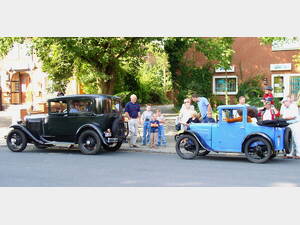 Zwei sehr alte Oldtimer fahren eine Straße entlang, Zuschauer beobachten sie vom Bürgersteig aus. Zwei sehr alte Oldtimer fahren eine Straße entlang, Zuschauer beobachten sie vom Bürgersteig aus.
