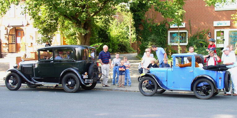 Zwei sehr alte Oldtimer fahren eine Straße entlang, Zuschauer beobachten sie vom Bürgersteig aus. Zwei sehr alte Oldtimer fahren eine Straße entlang, Zuschauer beobachten sie vom Bürgersteig aus.