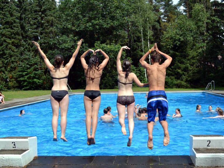 Drei Mädchen und ein Junge springen in ein Schwimmbecken. Im klaren Wasser sind weitere Schwimmerinnen und Schwimmer. Drei Mädchen und ein Junge springen in ein Schwimmbecken. Im klaren Wasser sind weitere Schwimmerinnen und Schwimmer.